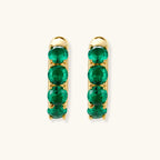 Close-up of gold-plated huggie hoop earrings featuring four emerald green cubic zirconia stones on white background.