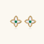 Gold quatrefoil stud earrings with white mother-of-pearl petals and emerald green gemstone center on beige background.