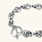 Silver stainless steel skull link bracelet with green crystal eye sockets and engraved detailing, toggle clasp closure.